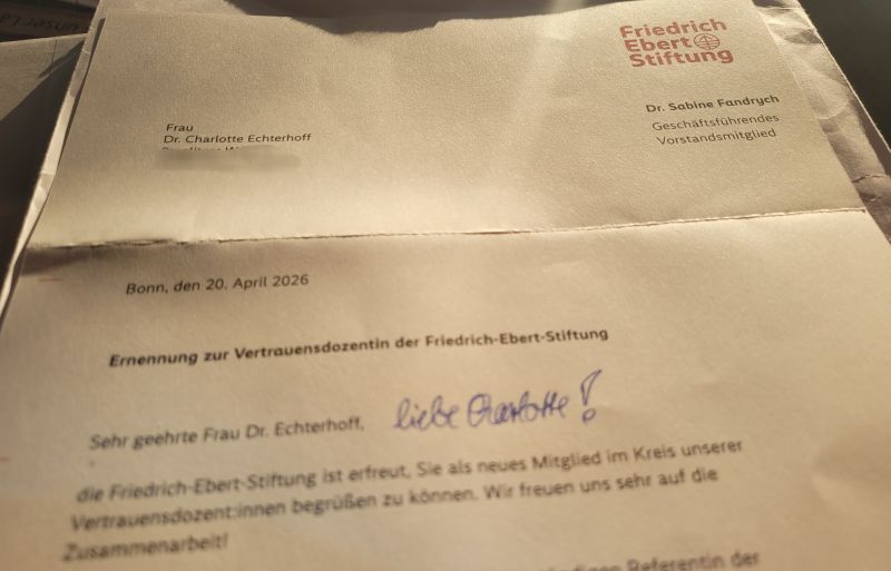 Dr. Charlotte Echterhoff hält einen Brief der Friedrich-Ebert-Stiftung in den Händen. Der Brief ist datiert auf den 20. April 2026 und richtet sich an sie als neue Vertrauensdozentin. Der Brief beginnt mit Sehr geehrte Frau Dr. Echterhoff, und enthält eine handschriftliche Notiz liebe Charlotte!. Im Hintergrund sind das Logo der Friedrich-Ebert-Stiftung und der Name von Dr. Sabine Fandrych zu sehen.