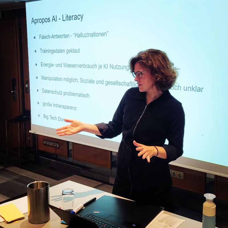Dr. Charlotte Echterhoff moderiert einen Workshop zum Thema **KI-Literacy (AI Literacy)**. Sie steht vor einer Präsentation mit der Überschrift *Apropos AI - Literacy* und erklärt mit gestenunterstrichener Klarheit die Herausforderungen von KI, wie **Falschantworten, Halluzinationen, Manipulationsmöglichkeiten und Datenschutzprobleme**. Auf dem Tisch vor ihr liegen ein Laptop, Notizen und eine Wasserflasche. Das Bild zeigt ihre **praktische und kritische Herangehensweise** an digitale Bildung.