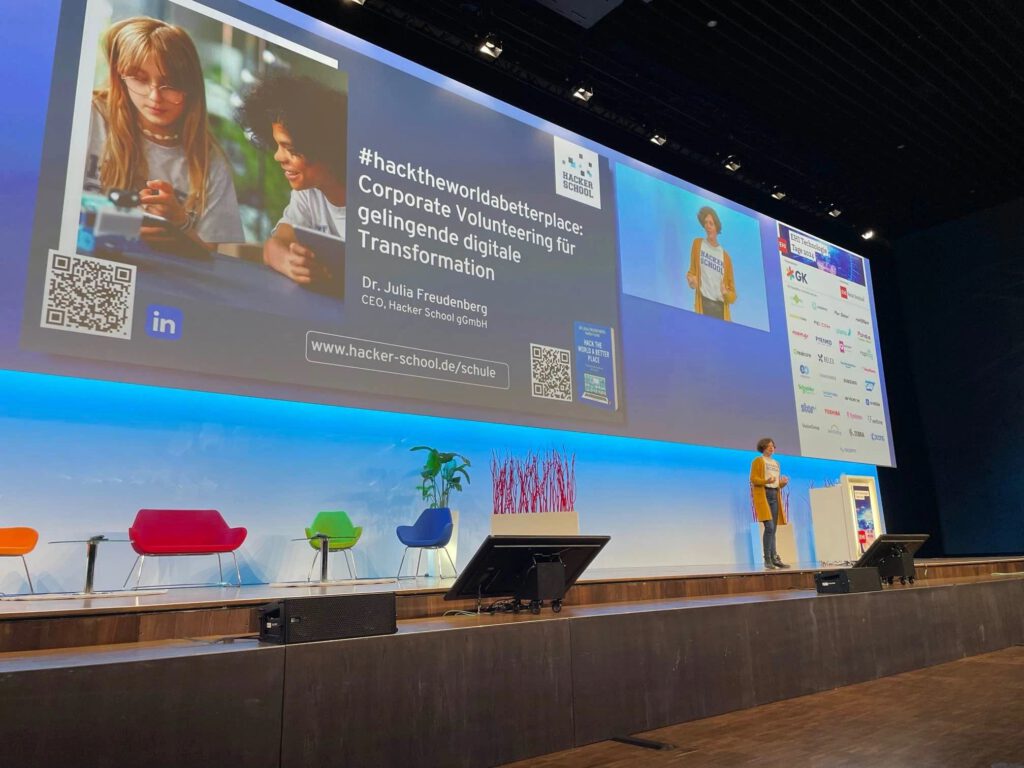 Dr. Charlotte Echterhoff steht auf einer großen Bühne und hält einen Vortrag. Im Hintergrund ist eine Leinwand mit der Präsentation von Dr. Julia Freudenberg, CEO der Hacker School gGmbH, zu sehen. Die Folie zeigt den Hashtag hacktheworldabetterplace und den Titel Corporate Volunteering für gelingende digitale Transformation. Auf der Bühne stehen farbige Sessel, eine Pflanze und ein Rednerpult. Die Veranstaltung findet auf der EHI Tech Summit 2024 statt.