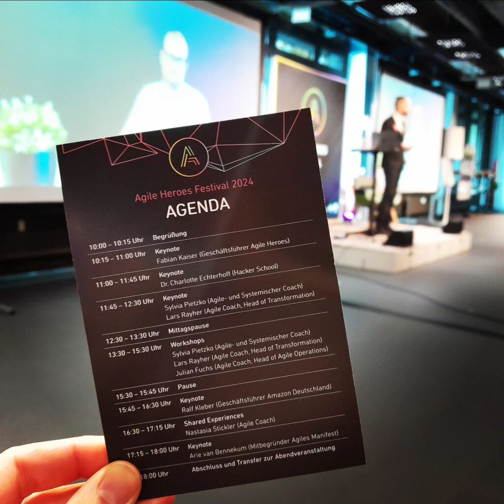 Dr. Charlotte Echterhoff ist als Rednerin auf dem Agile Heroes Festival 2024 gelistet. Die Agenda zeigt, dass sie von 11:00 bis 11:45 Uhr eine Keynote hält. Im Hintergrund ist eine Bühne mit einer Person zu sehen, die eine Präsentation hält.