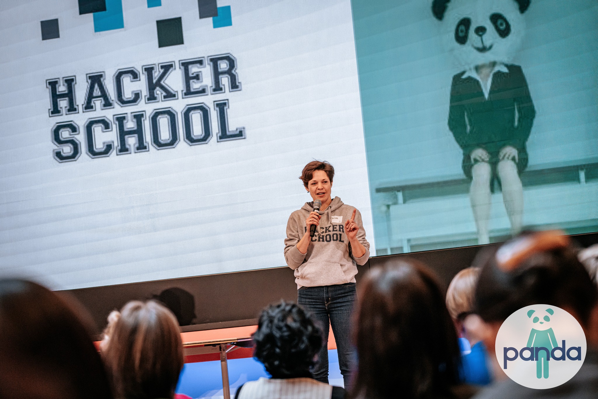 Dr. Charlotte Echterhoff hält einen Vortrag auf der PANDA Change & Innovation 2023. Sie steht auf einer Bühne vor einem großen Bildschirm mit dem Hacker School Logo und einem Panda-Maskottchen in Anzug. Sie trägt ein graues Hacker School Hoodie, Jeans und hält ein Mikrofon in der Hand. Im Vordergrund sind die Köpfe von Zuhörern zu sehen.