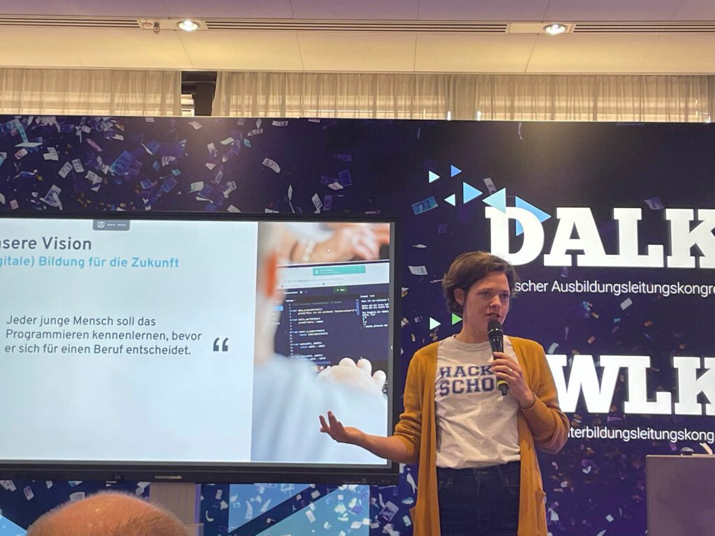 Dr. Charlotte Echterhoff hält einen **Vortrag auf dem Deutschen Ausbildungskongress (DALK/WLK)** zum Thema *digitale Bildung für die Zukunft*. Sie steht mit Mikrofon in der Hand vor einem großen Bildschirm, der ihr Zitat *‚Jeder junge Mensch soll das Programmieren kennenlernen, bevor er sich für einen Beruf entscheidet.‘* zeigt. Im Hintergrund ist ein **Bühnenbanner mit dem Logo der Konferenz** zu sehen. Das Bild unterstreicht ihre **engagierte und visionäre Art**, digitale Kompetenzen in der Bildung zu verankern.