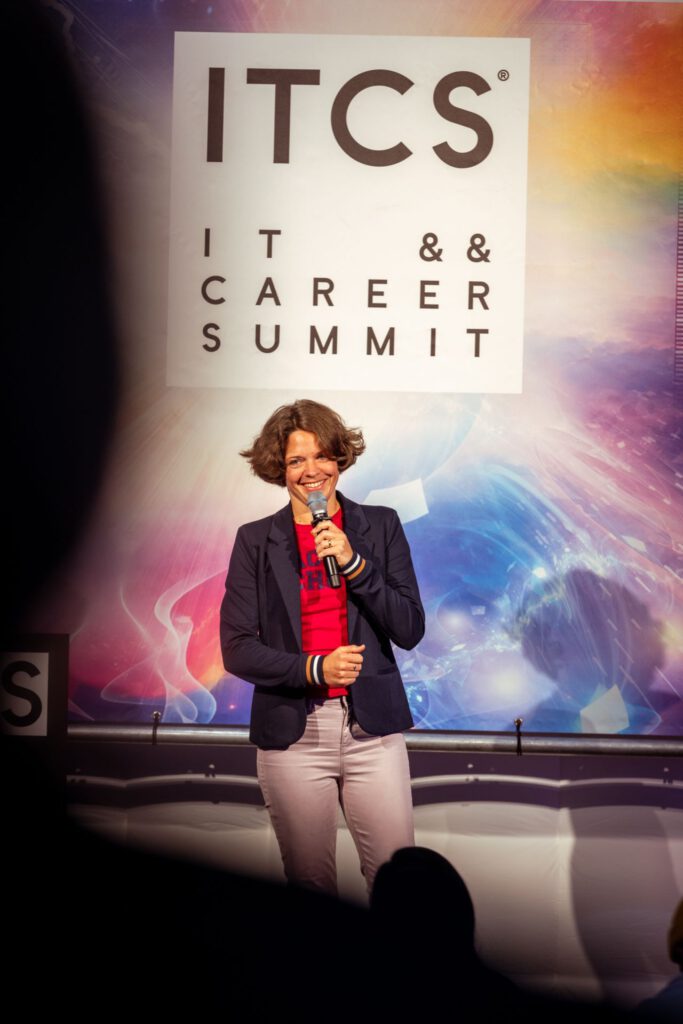 Dr. Charlotte Echterhoff hält einen Vortrag auf dem ITCS IT & Career Summit. Sie steht auf einer Bühne vor einem großen Banner mit der Aufschrift ITCS IT & Career Summit. Sie trägt eine dunkle Blazerjacke, ein rotes T-Shirt und helle Hosen. In der Hand hält sie ein Mikrofon.