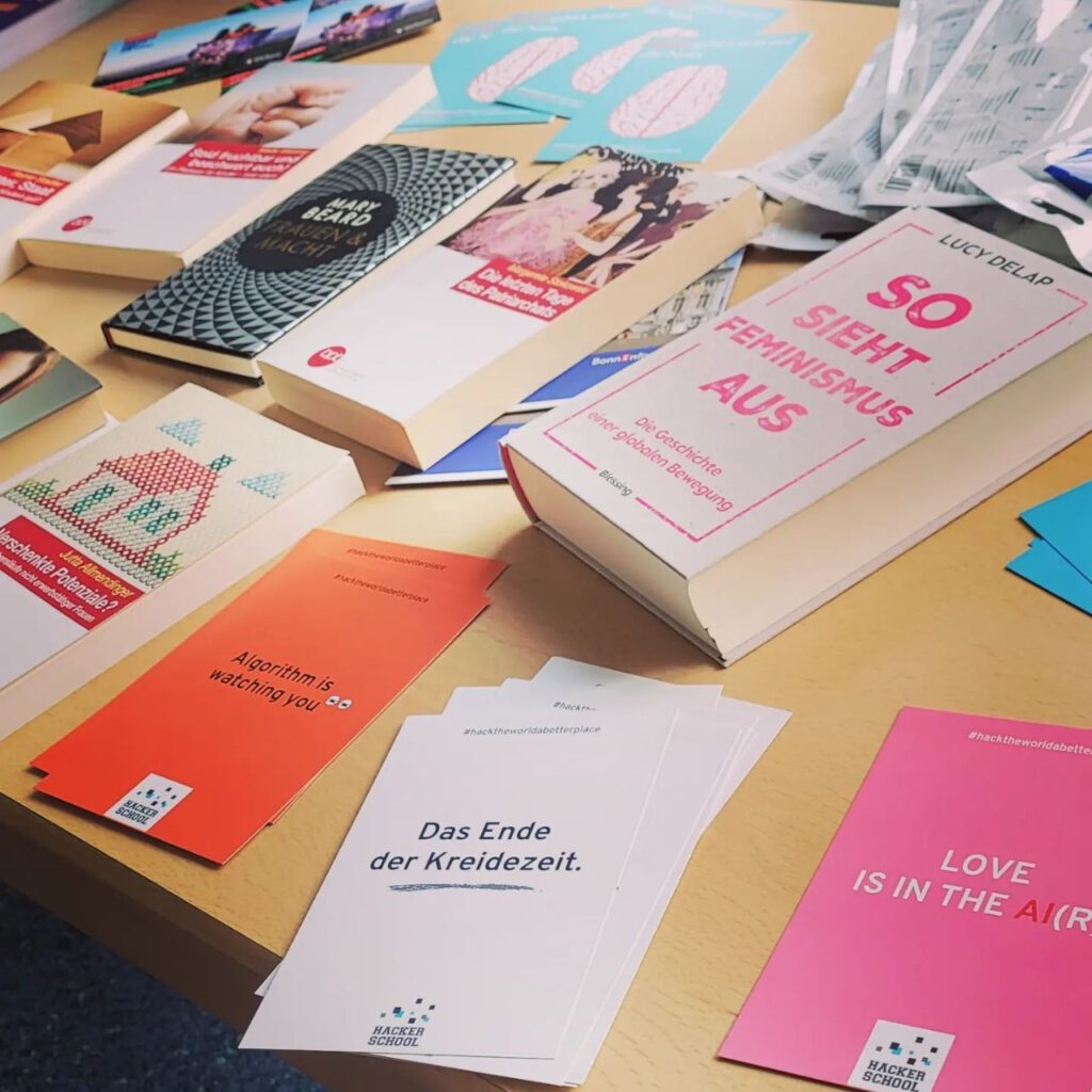 Dr. Charlotte Echterhoffs Auslage mit verschiedenen Büchern und Materialien. Zu sehen sind Bücher wie So sieht Feminismus aus von Lucy Delap, Frauen und Macht von Mary Beard, Die letzten Tage des Patriarchats von Margarete Stochowski, sowie Flyer und Karten mit Aufschriften wie Algorithm is watching you, Das Ende der Kreidezeit und LOVE IS IN THE AI. Die Materialien liegen auf einem Holztisch und sind teilweise mit dem Hashtag hacktheworldabetterplace der Hacker School versehen.