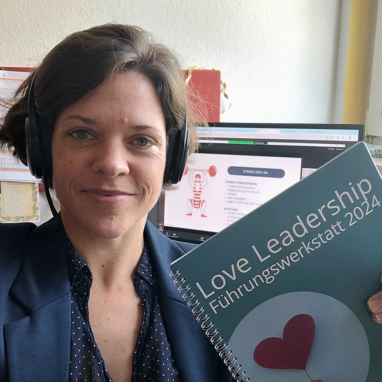 Dr. Charlotte Echterhoff hält ein Notizbuch mit der Aufschrift Love Leadership Führungswerkstatt 2024 in der Hand. Sie trägt ein dunkles Blazerjackett über einem gepunkteten Oberteil und hat ein Headset auf. Im Hintergrund ist ein Laptop mit einer Präsentation zu sehen, auf der ein stilisiertes Bild einer Person mit einer Hantel und der Text STRENG DICH AN zu erkennen ist.