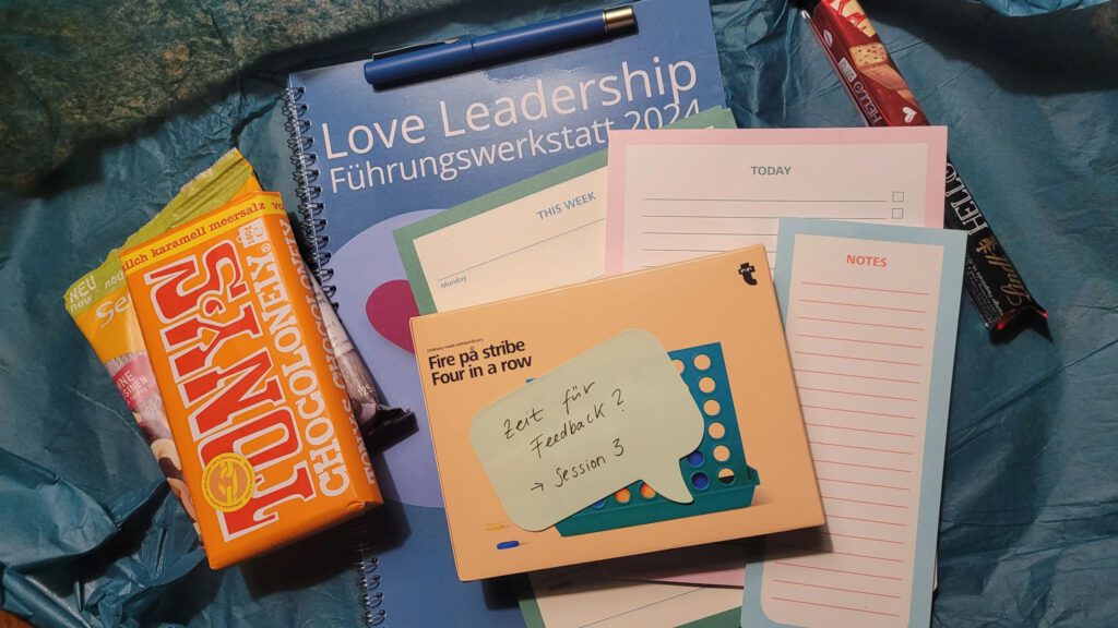 Auf dem Bild liegen verschiedene Materialien der Love Leadership Führungswerkstatt 2024 von Dr. Charlotte Echterhoff. Dazu gehören ein Notizbuch mit der Aufschrift Love Leadership Führungswerkstatt 2024, ein Heft mit der Beschriftung Fire på stripe / Four in a row, ein Notizblock mit der handschriftlichen Notiz "Zeit für Feedback? → Session 3", ein leerer Wochenplaner sowie Snacks wie eine Packung Tony’s Chocolonely und eine Packung Kekse. Alles liegt auf einer blauen Unterlage.