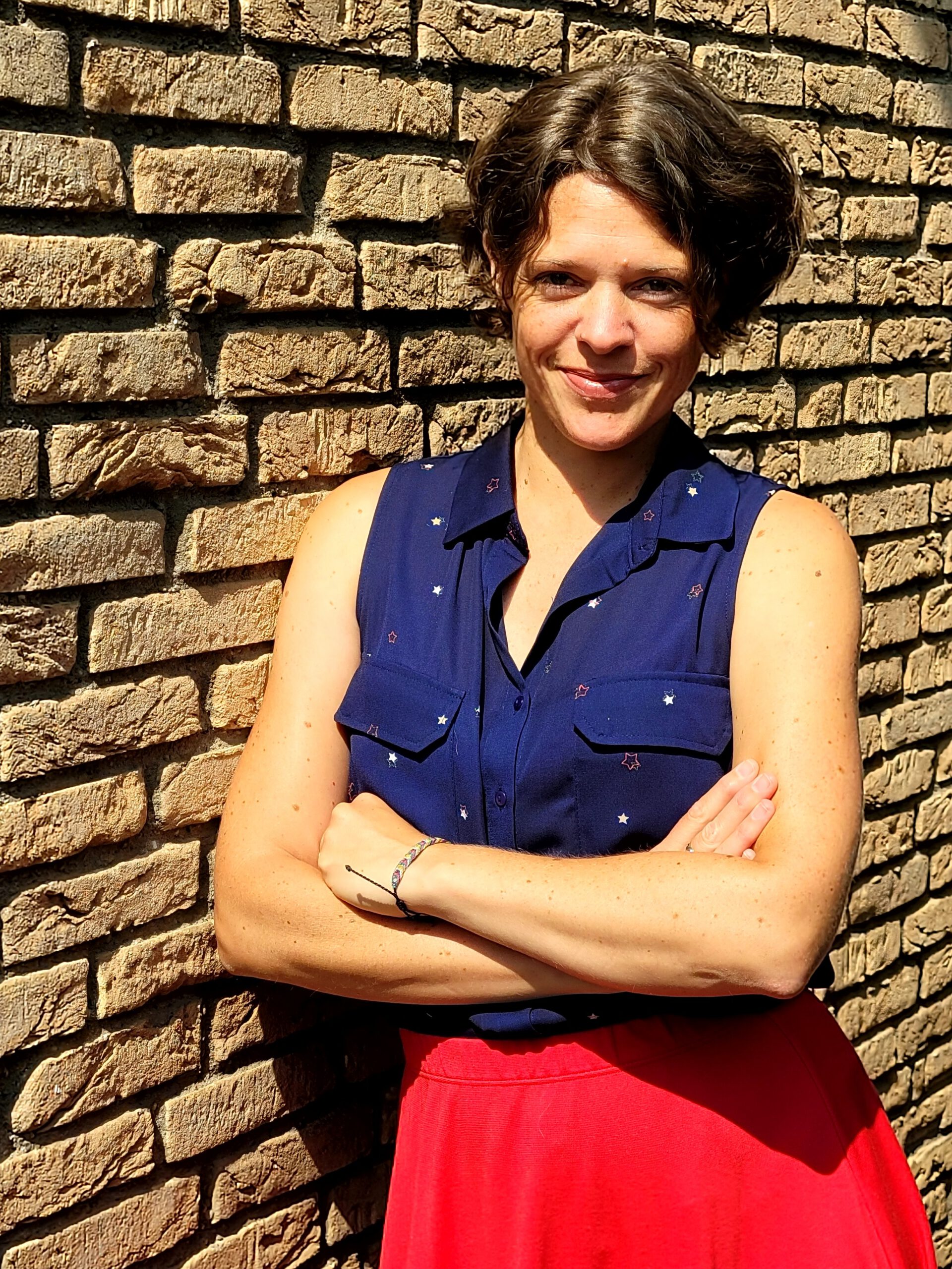 Dr. Charlotte Echterhoff steht lächelnd mit verschränkten Armen vor einer Steinmauer. Sie trägt ein dunkles, kurzärmliges Oberteil mit Sternenmuster und einen roten Rock. Ihr Haar ist kurz geschnitten, und sie wirkt selbstbewusst und entspannt.
