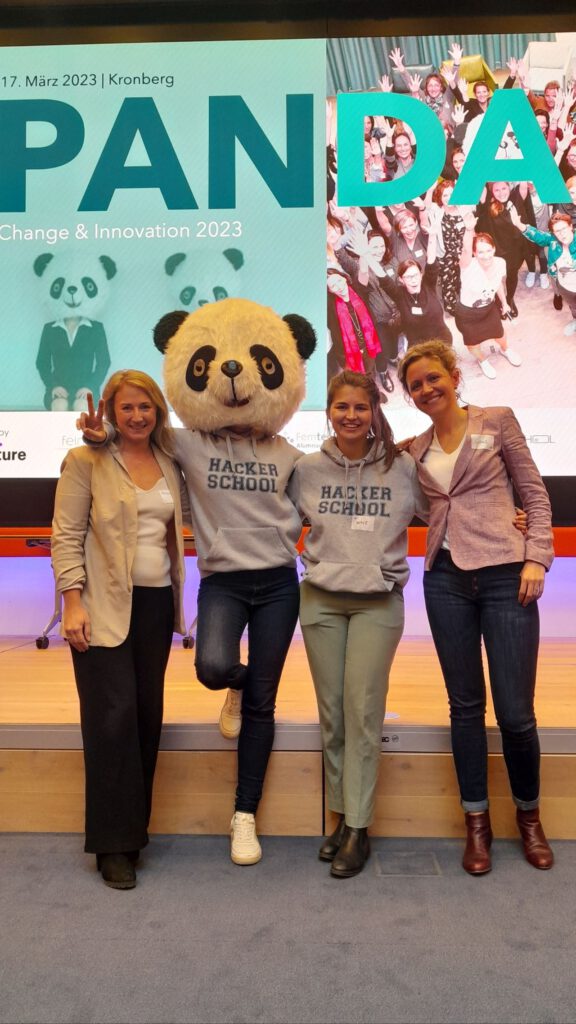 Dr. Charlotte Echterhoff steht in der Mitte einer Gruppe von vier Personen auf der PANDA Change & Innovation 2023 in Kronberg. Sie trägt ein graues Hacker School Hoodie und ein Panda-Kostüm. Die anderen tragen ebenfalls Hoodies oder Blazer und lächeln in die Kamera.