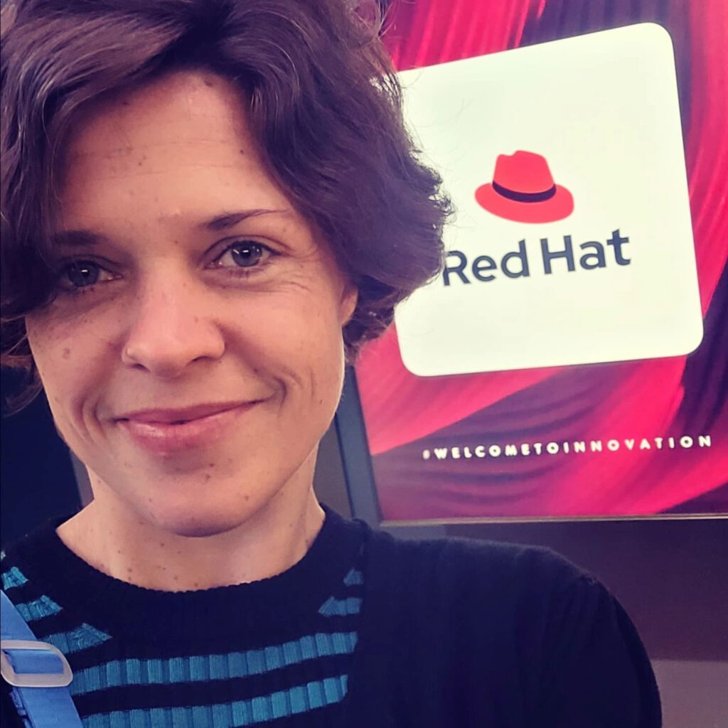 Dr. Charlotte Echterhoff vor einem Red Hat Stand mit dem Slogan #WELCOMETOINNOVATION. Sie trägt ein schwarzes Oberteil mit blauen Streifen und einen blauen Gurt. Im Hintergrund ist das Red Hat Logo mit einem roten Hut zu sehen.